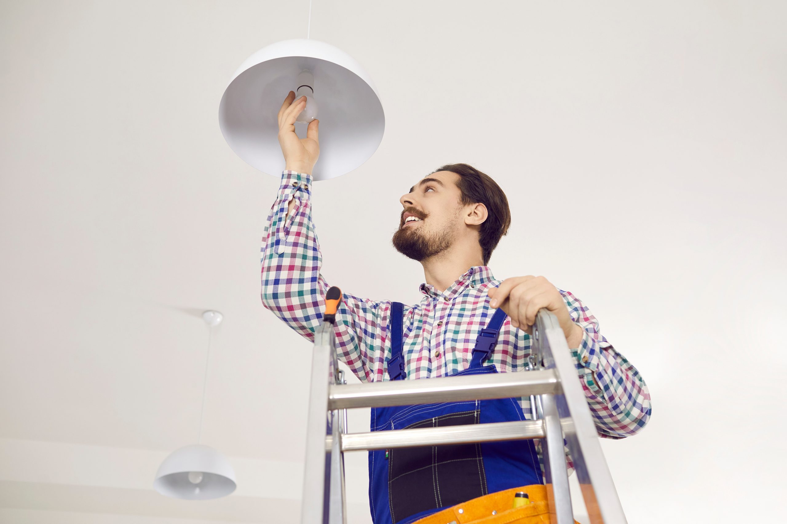 Electrician screwing in light bulb at home. Happy man in work uniform standing on ladder in residential building, office, school or hospital and changing LED lightbulb in modern white lamp on ceiling